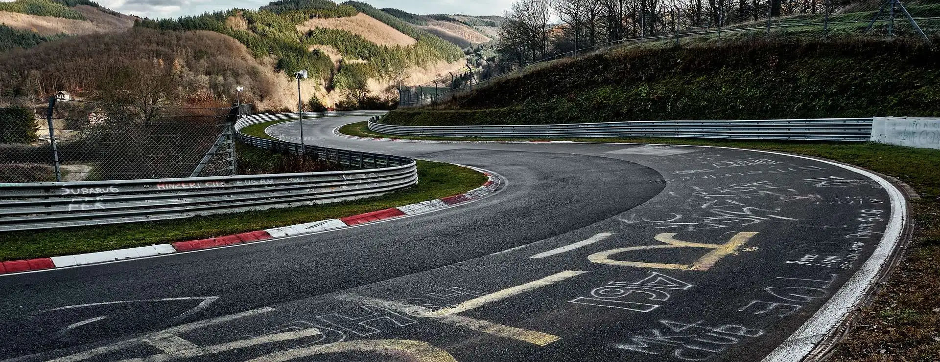Abgenutzte Asphalt-Rennstrecke mit roten und weißen Bordsteinen, Graffiti-Markierungen und Metall-Leitplanken.