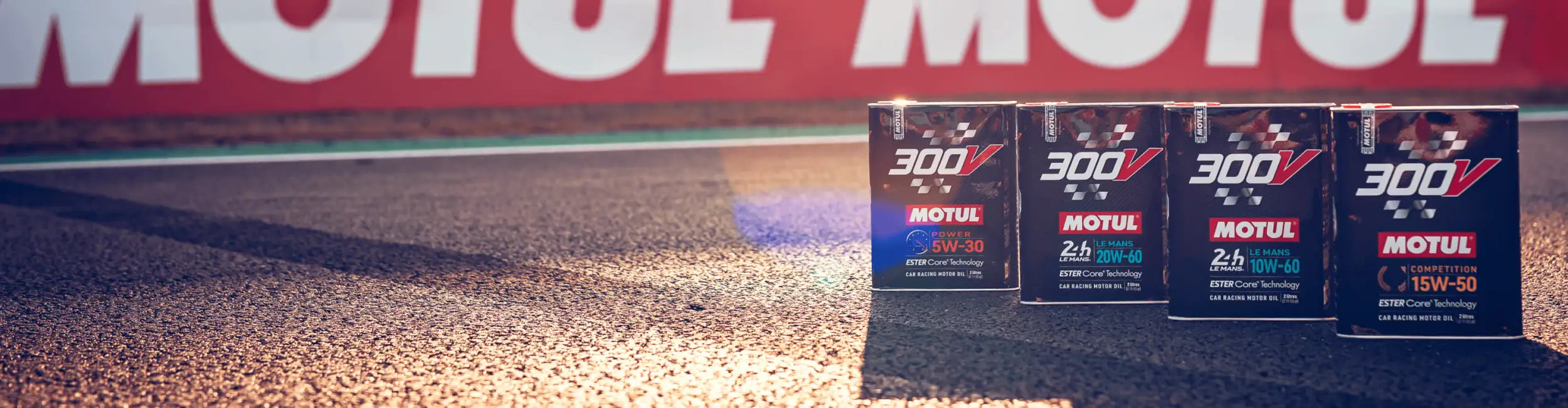 Vier schwarze Motorölkanister Motul 300v mit roten und blauen Etiketten, in einer Reihe auf einer Asphaltbahn angeordnet.